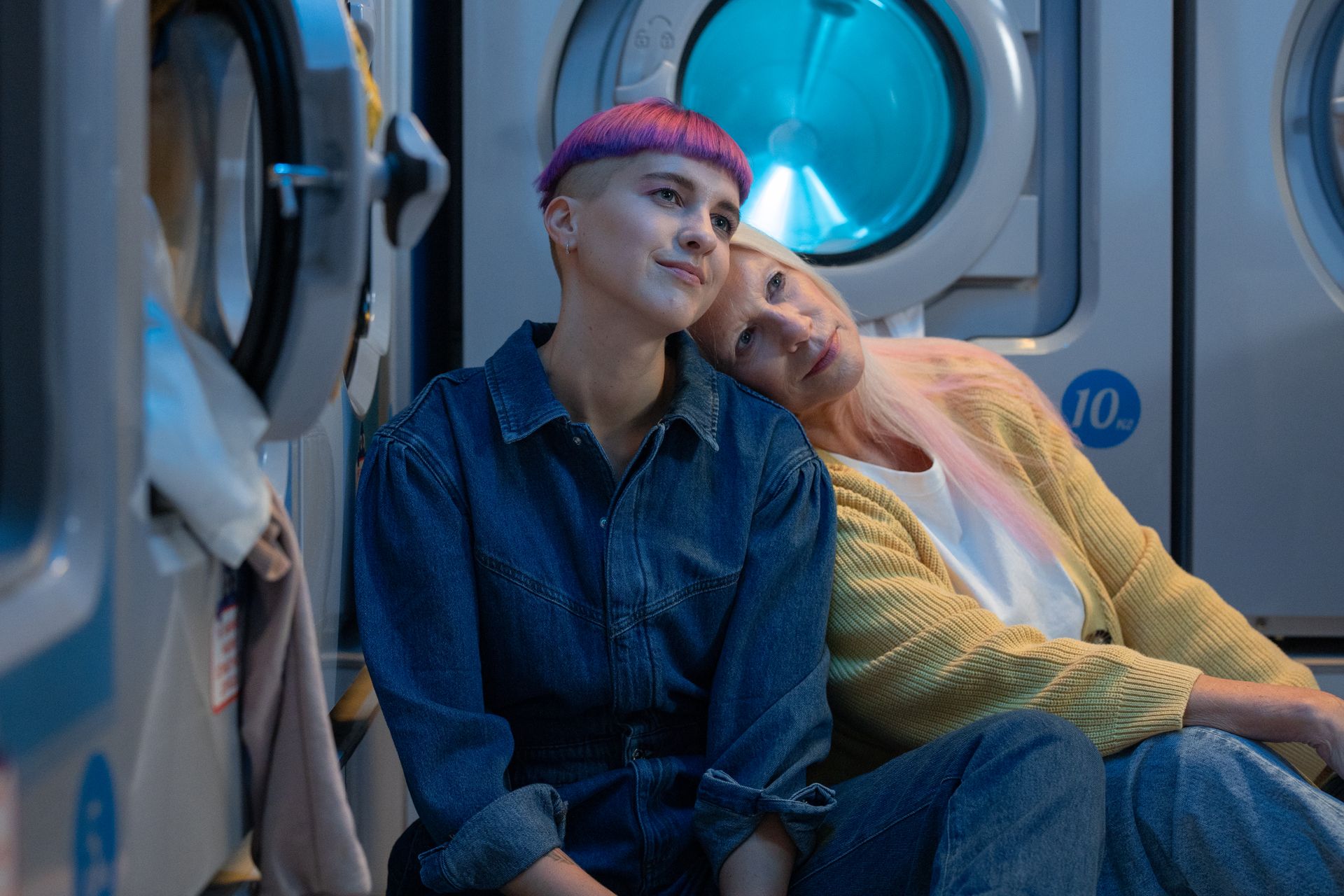 Two women share a tender moment sitting beside washing machines in a laundromat.
