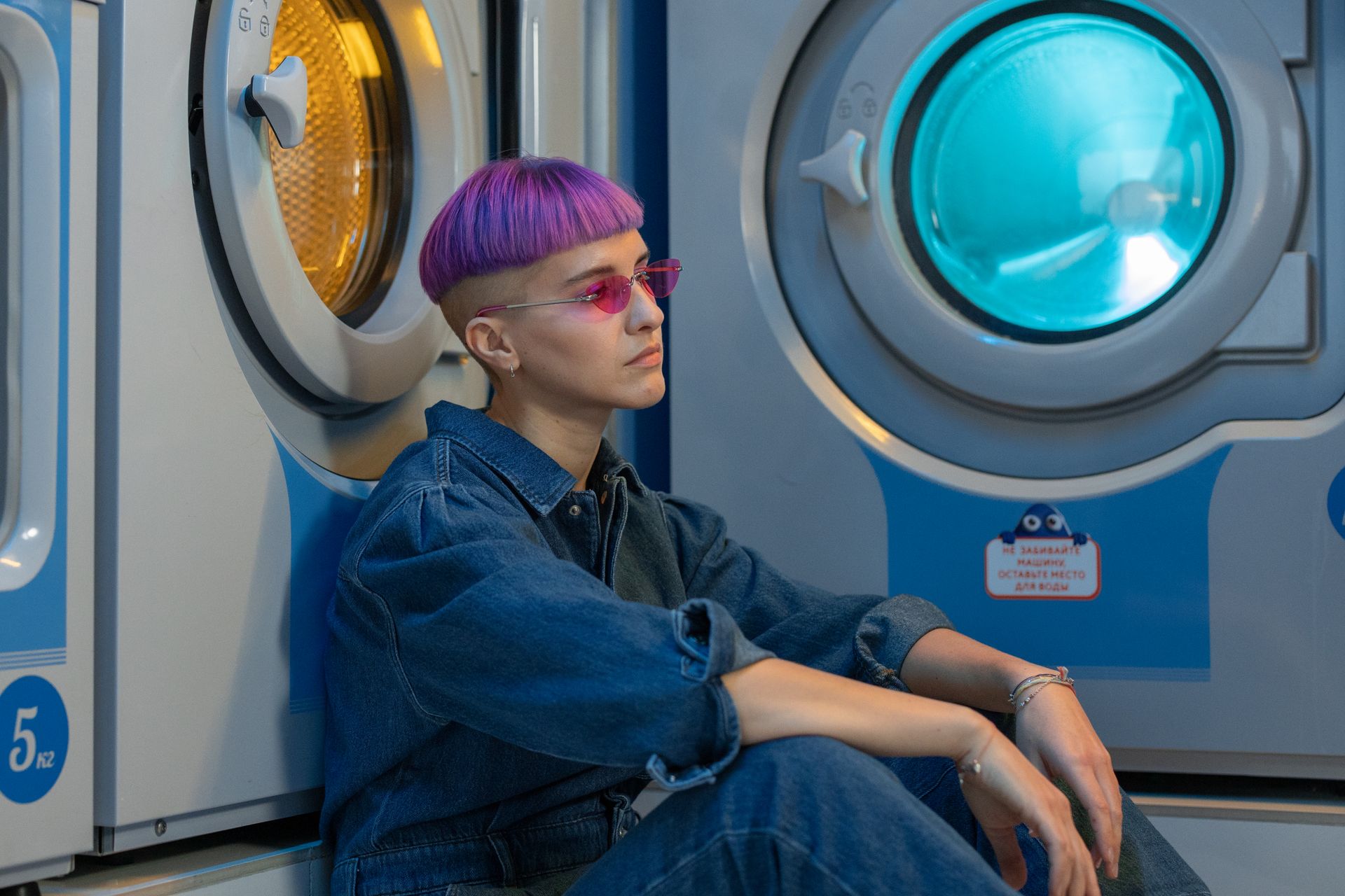 A stylish person with purple hair sits by washing machines in a modern laundry facility.