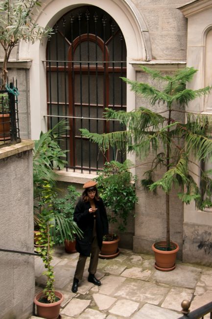 Full length of person in warm clothes and hat standing in courtyard on pavement near green potted plants and building with arched window in daylight