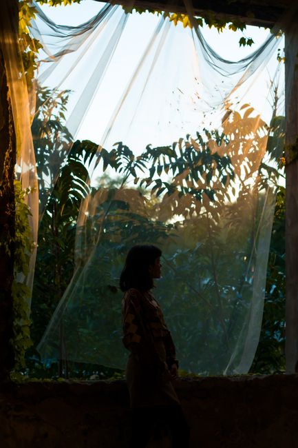Side view of anonymous woman standing in dark building near window with textile and green plants with foliage in evening