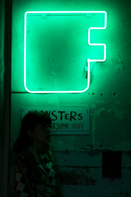 Female leaning on wall of building with green neon signboard in dark street