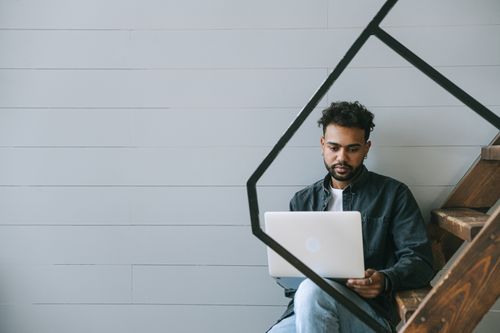 Free Man sitting on stairs working on laptop in a minimalist setting, reflecting modern work-from-home lifestyle. Stock Photo