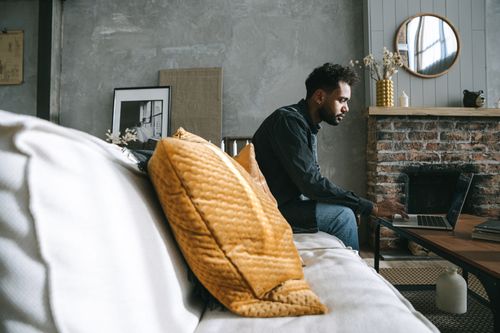 Free A man sitting on a couch using a laptop in a stylish living room, reflecting remote work culture. Stock Photo