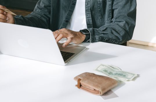 Close-up of a man using a laptop for online purchases, featuring a wallet and cash on the table.