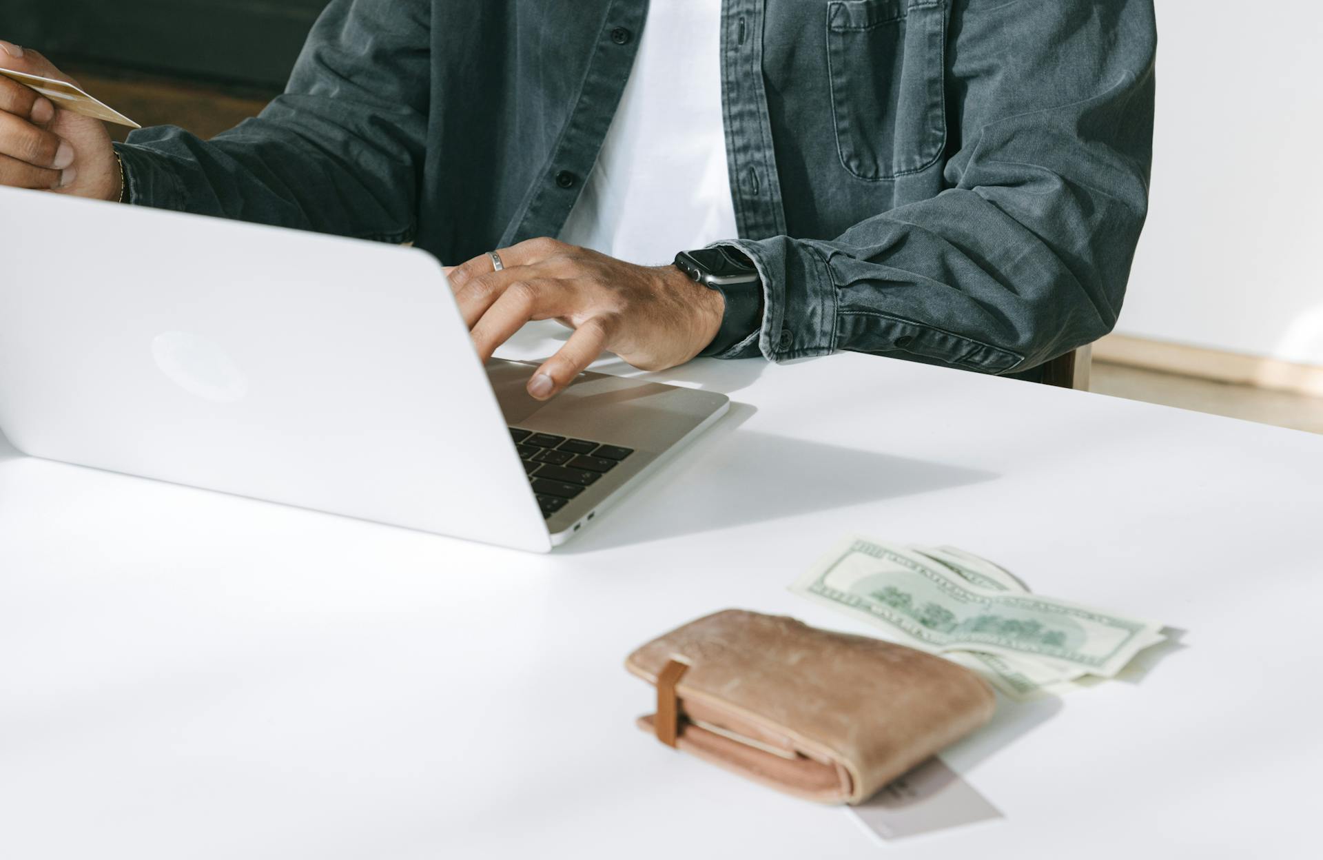 Close-up of a man using a laptop for online purchases, featuring a wallet and cash on the table.