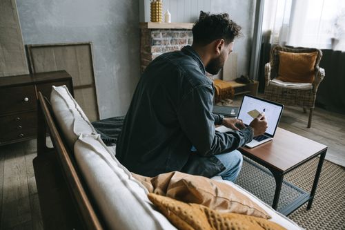 Free A man wearing a denim jacket makes an online purchase using his laptop while sitting indoors on a couch. Stock Photo