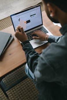 A person using a laptop for online shopping with a card on a wooden table.