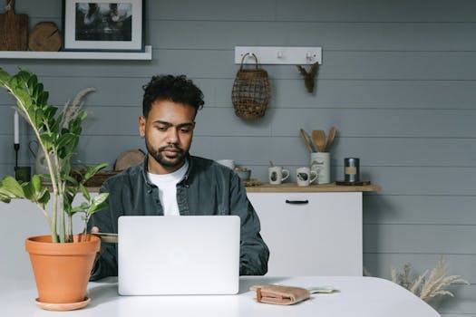 Adult man focused on laptop work, sitting at home with a potted plant on the table.