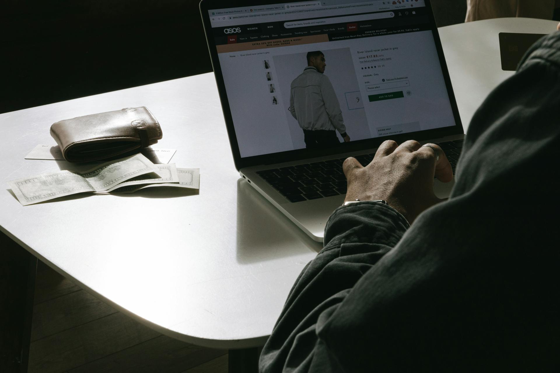 Adult man shopping for clothes online at home using a laptop, with a wallet on the table.