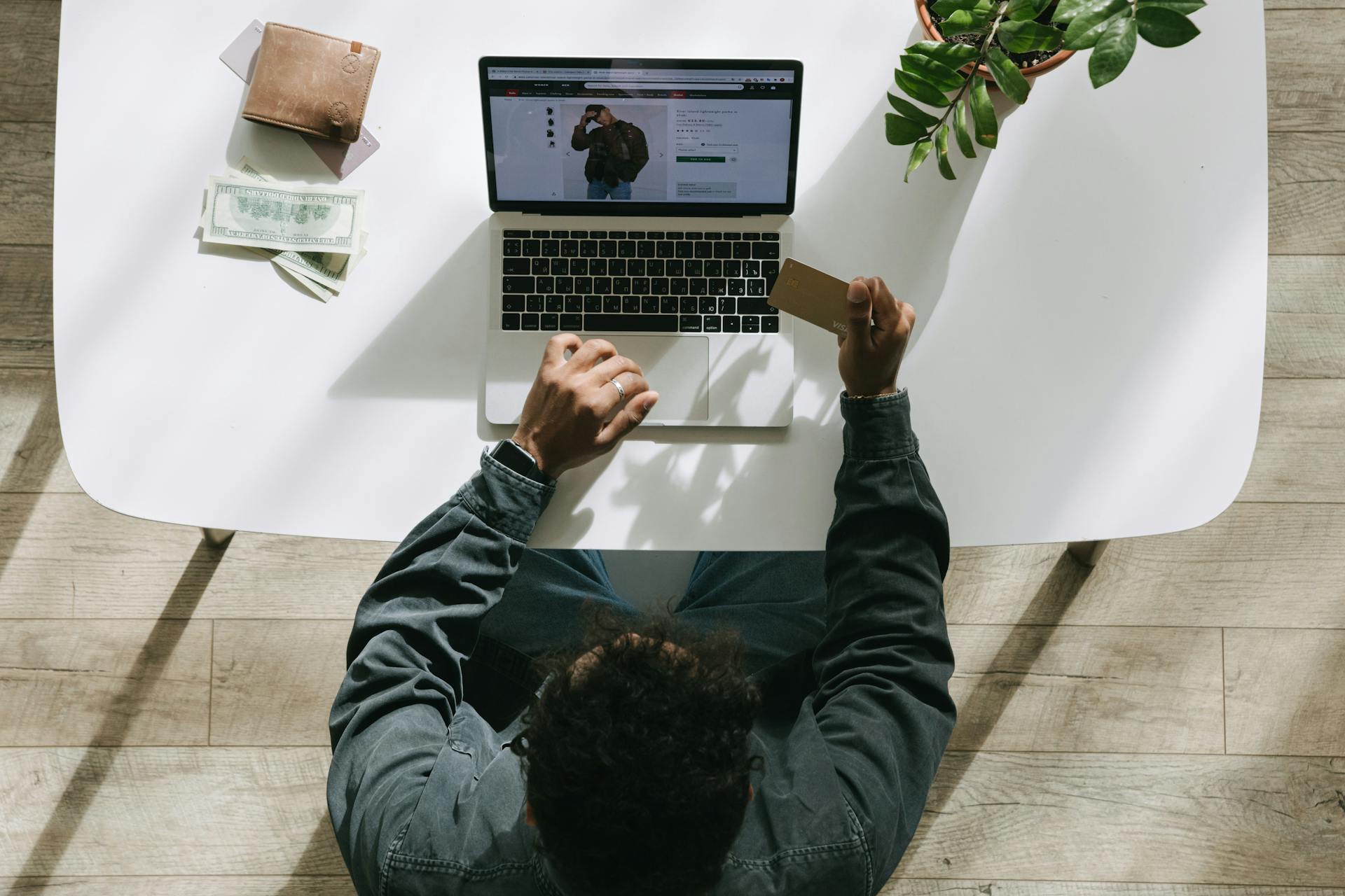 Person using a laptop for online shopping, holding credit card at a bright indoor desk.