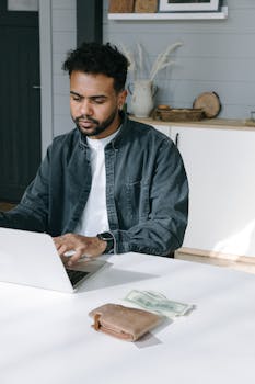 Man working remotely on laptop at home with paper money and wallet on white table.