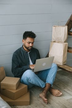 Young man using a laptop surrounded by shopping bags and packages, working on eCommerce business.