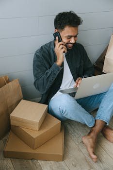 Young man sitting on floor multitasking with packages, laptop, and phone.