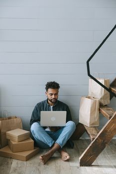 Man sitting on floor with laptop surrounded by shopping bags and boxes, enjoying online shopping experience.