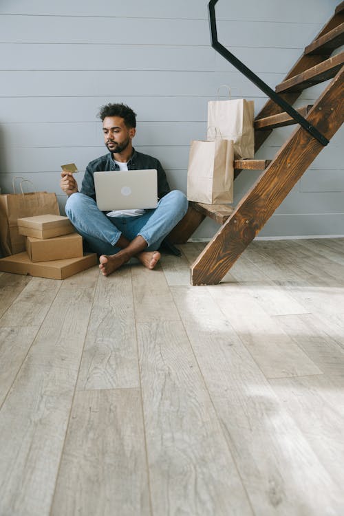 A Man Sitting on the Wooden Flooring while Looking at His Debit Card