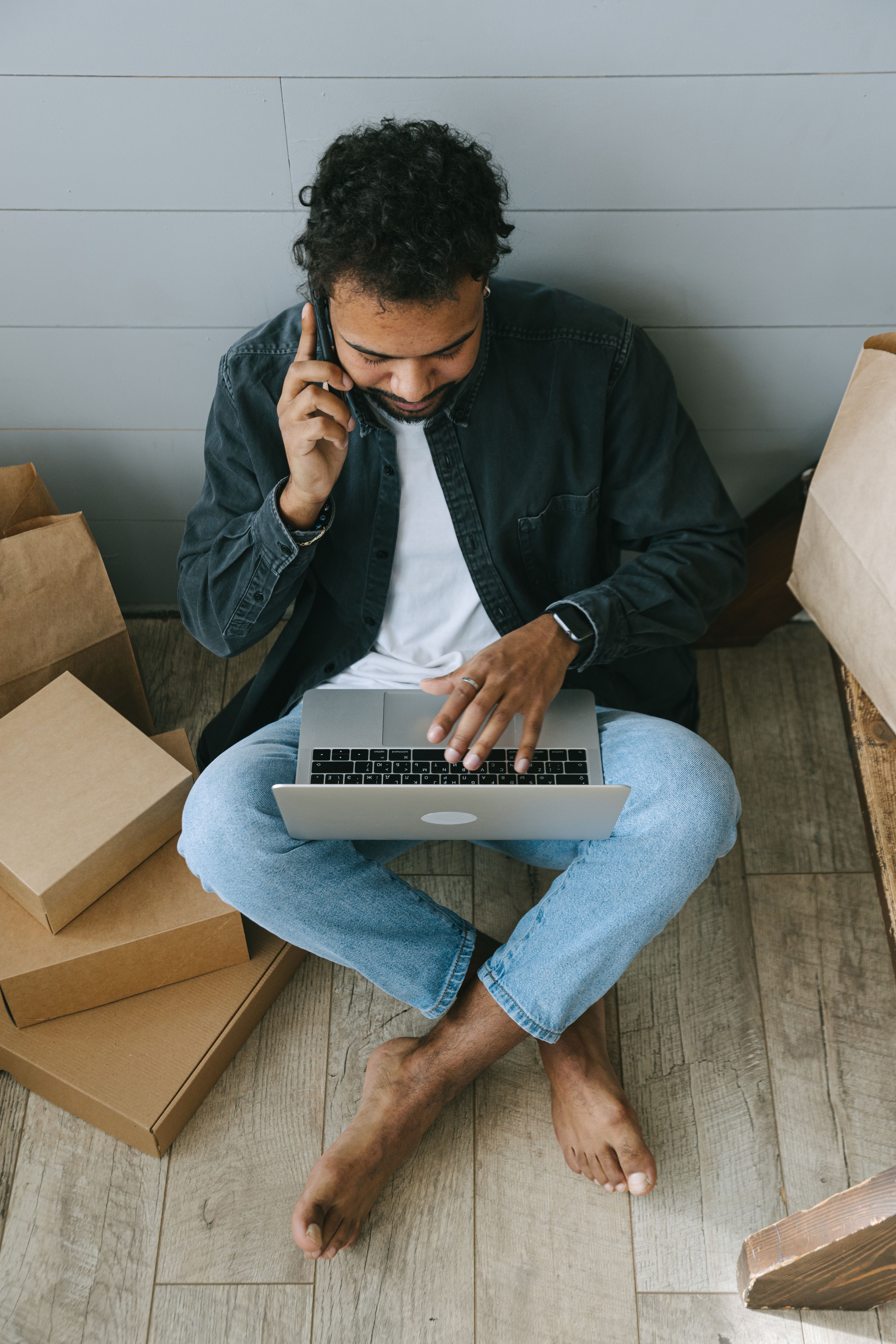 Man sitting barefoot on wooden floor using laptop and talking on phone surrounded by boxes.