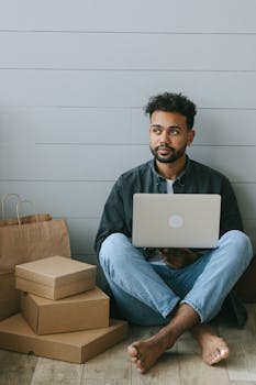 Barefoot man sits with laptop and packages, embodying modern remote work lifestyle.