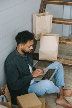 Young man sits barefoot on wooden floor working on laptop surrounded by paper shopping bags.