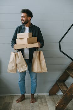 A smiling man holding boxes and paper bags, standing near stairs indoors.