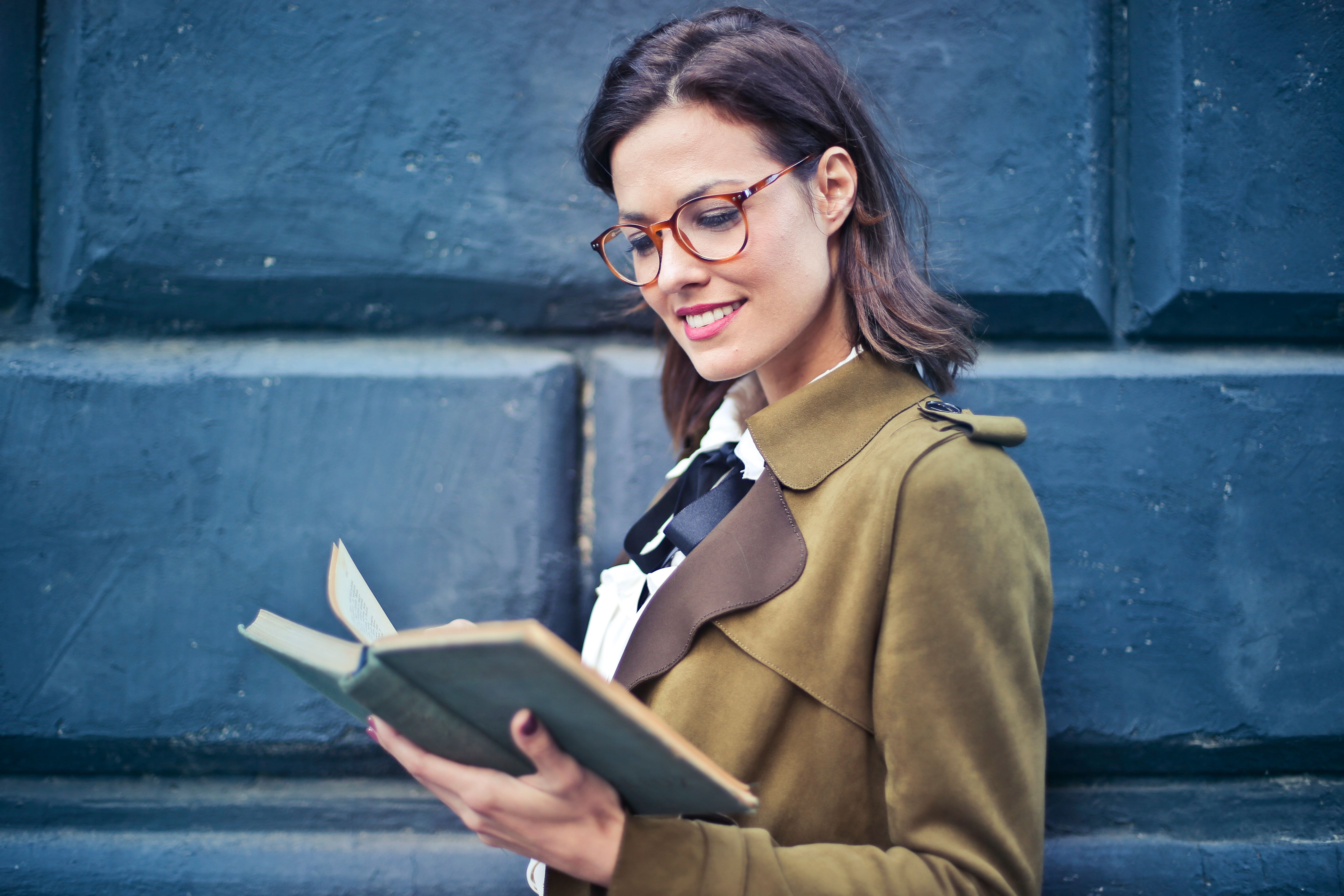 Smiling woman enjoying a book while standing against a textured blue wall