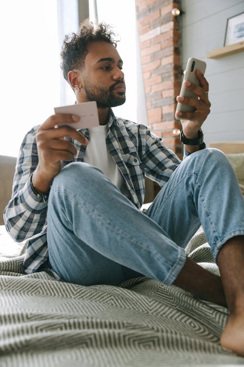 A Man in Plaid Long Sleeves Sitting on the Bed while Holding a Cellphone and Card