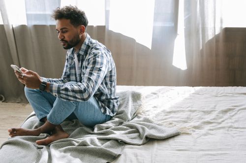 Free A bearded man in denim and plaid sits barefoot on bed using smartphone Stock Photo