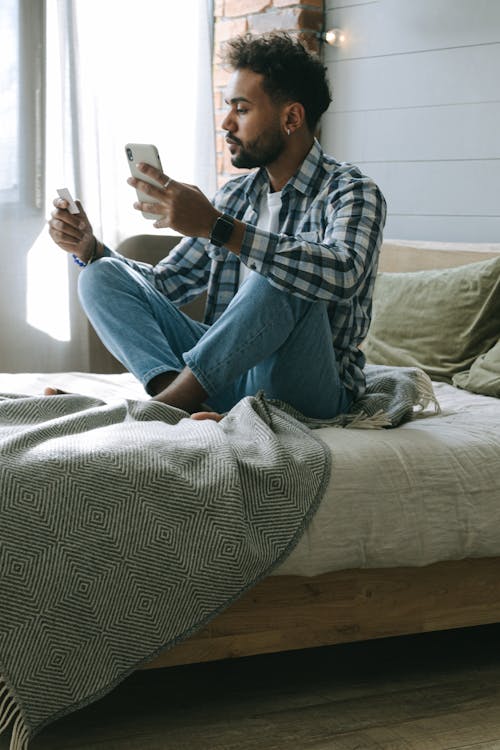 A Man in Plaid Long Sleeves Sitting on the Bed while Busy Looking at the Visa Card he is Holding