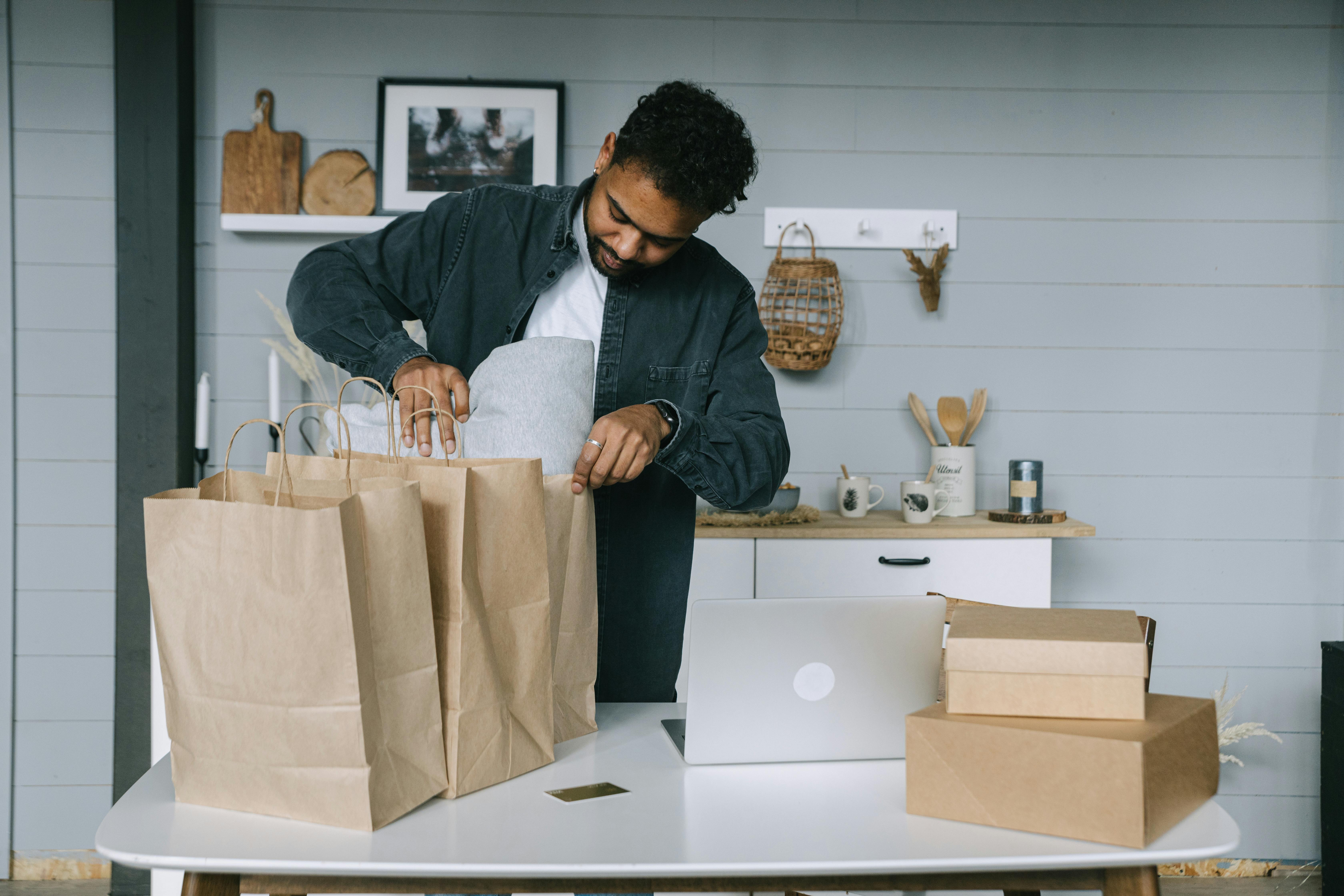 Young man organizing paper bags near a laptop in a cozy indoor setting.
