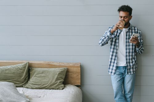 Free Young man enjoys coffee and checks phone, leaning against wall in cozy bedroom. Stock Photo