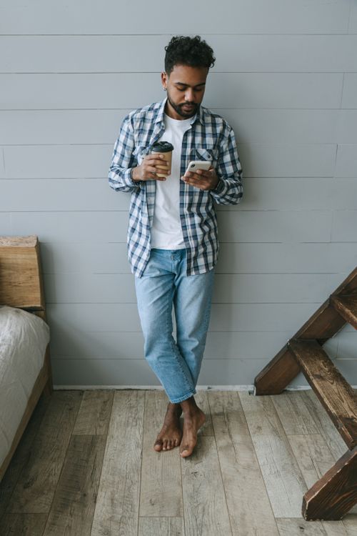 Free Bearded man in casual attire standing indoors holding a cup and using a smartphone. Stock Photo