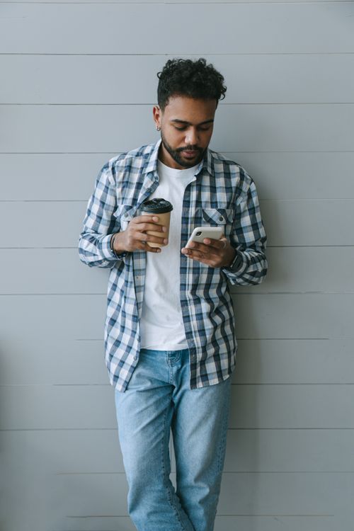 Free Casual young man in a plaid shirt enjoying coffee while browsing on his smartphone indoors. Stock Photo