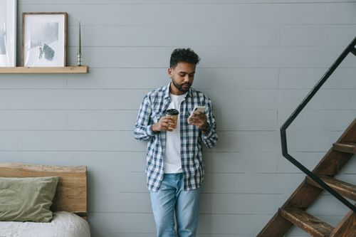 Free A man in a plaid shirt stands indoors, holding coffee and using a smartphone. Stock Photo