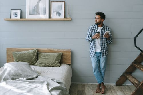 Free A relaxed man leans against a wall holding coffee and phone in a cozy bedroom. Stock Photo
