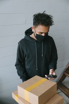 Courier wearing a face mask handles cardboard packages and smartphone indoors.