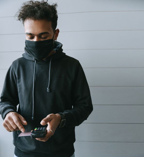 A man wearing a face mask processes a contactless payment indoors with a card and terminal. A man wearing a face mask processes a contactless payment indoors with a card and terminal.