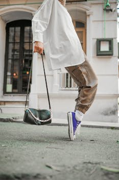 A fashionable person in urban setting wearing sneakers, brown pants, and white blouse, carrying a stylish bag.
