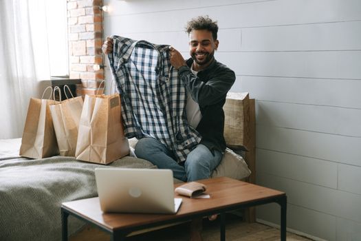 Smiling man excitedly unpacking new clothes from shopping bags in his cozy bedroom setting.