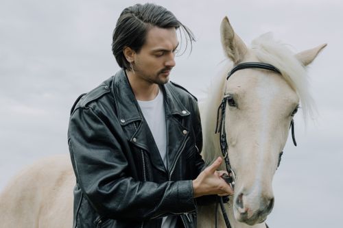 Free Man wearing a black leather jacket tenderly holding a white horse outdoors. Stock Photo
