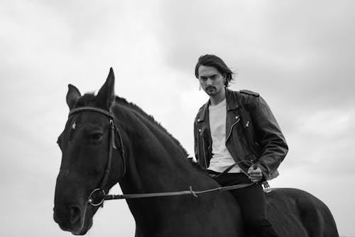 Grayscale Photo of a Man in Black Leather Jacket Riding a Horse