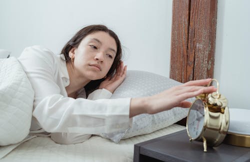 Free Sleepy young brunette female in white sleepwear resting in comfortable bed and waking up with alarm clock at home in morning time Stock Photo