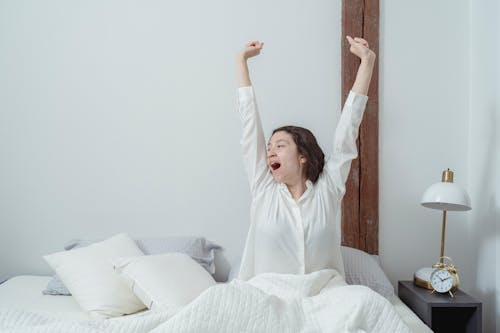 Sleepy brunette sitting on white comfortable bed under blanket and stretching arms up while yawning and looking away during morning time at home