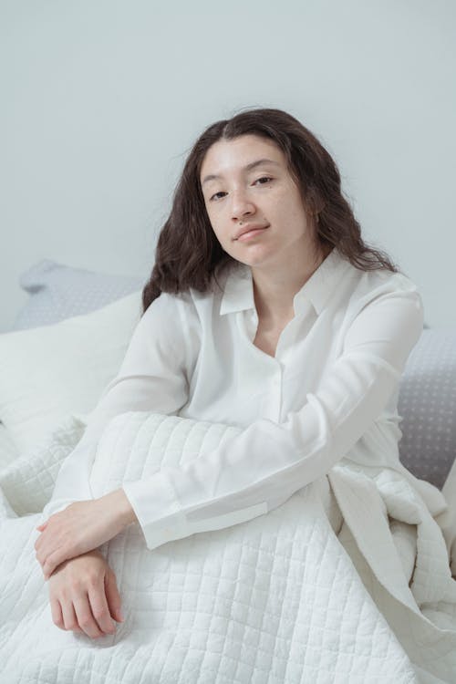 Charming young brunette in white nightwear sitting on comfortable bed under white blanket embracing knees and looking at camera against white background