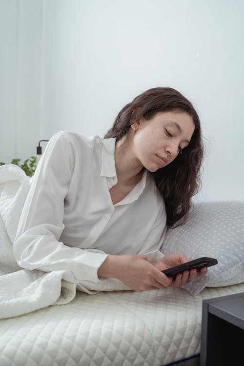 Free Young brunette in white sleepwear relaxing on comfortable bed under white blanket leaning on elbow and using mobile phone in morning time at home Stock Photo