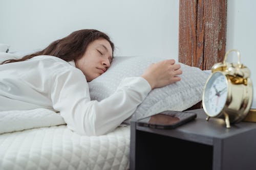 Free Young woman with dark long wavy hair sleeping peacefully on belly on comfortable bed under white blanket near bedside table with alarm clock and smartphone Stock Photo