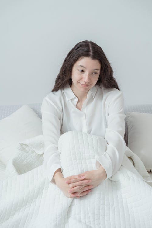 From above of charming young female with long dark wavy hair and in white sleepwear sitting on comfortable bed under blanket embracing knees and looking away against white wall