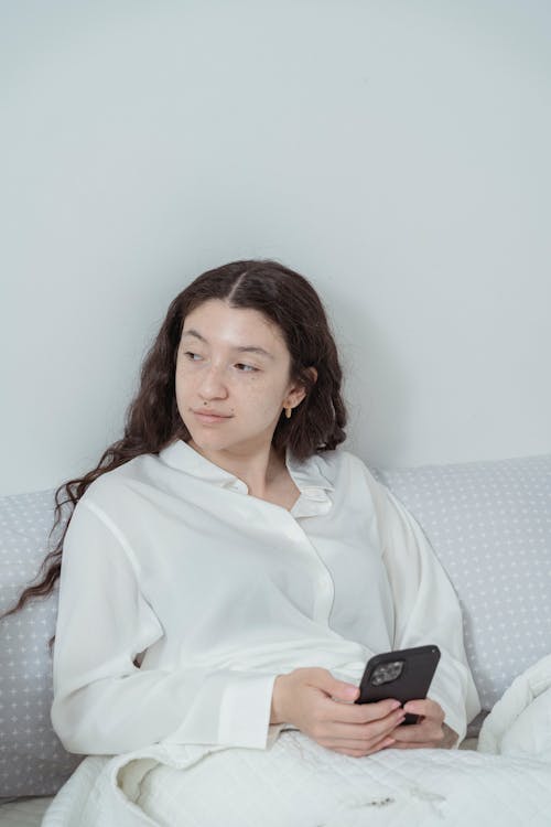 Young woman with long dark wavy hair in white sleepwear sitting on comfortable white bed and using smartphone while looking away against white wall in morning time