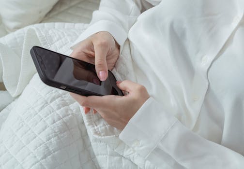 Close-up of a woman's hands holding a smartphone while relaxing indoors, wearing a white shirt.