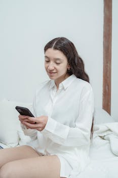A woman with long hair sits on a bed, smiling while texting on her smartphone in a bright room.