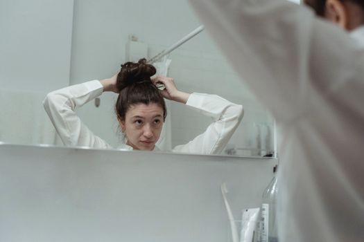 Young woman styling her hair in a bathroom mirror reflection, creating a casual and serene moment.