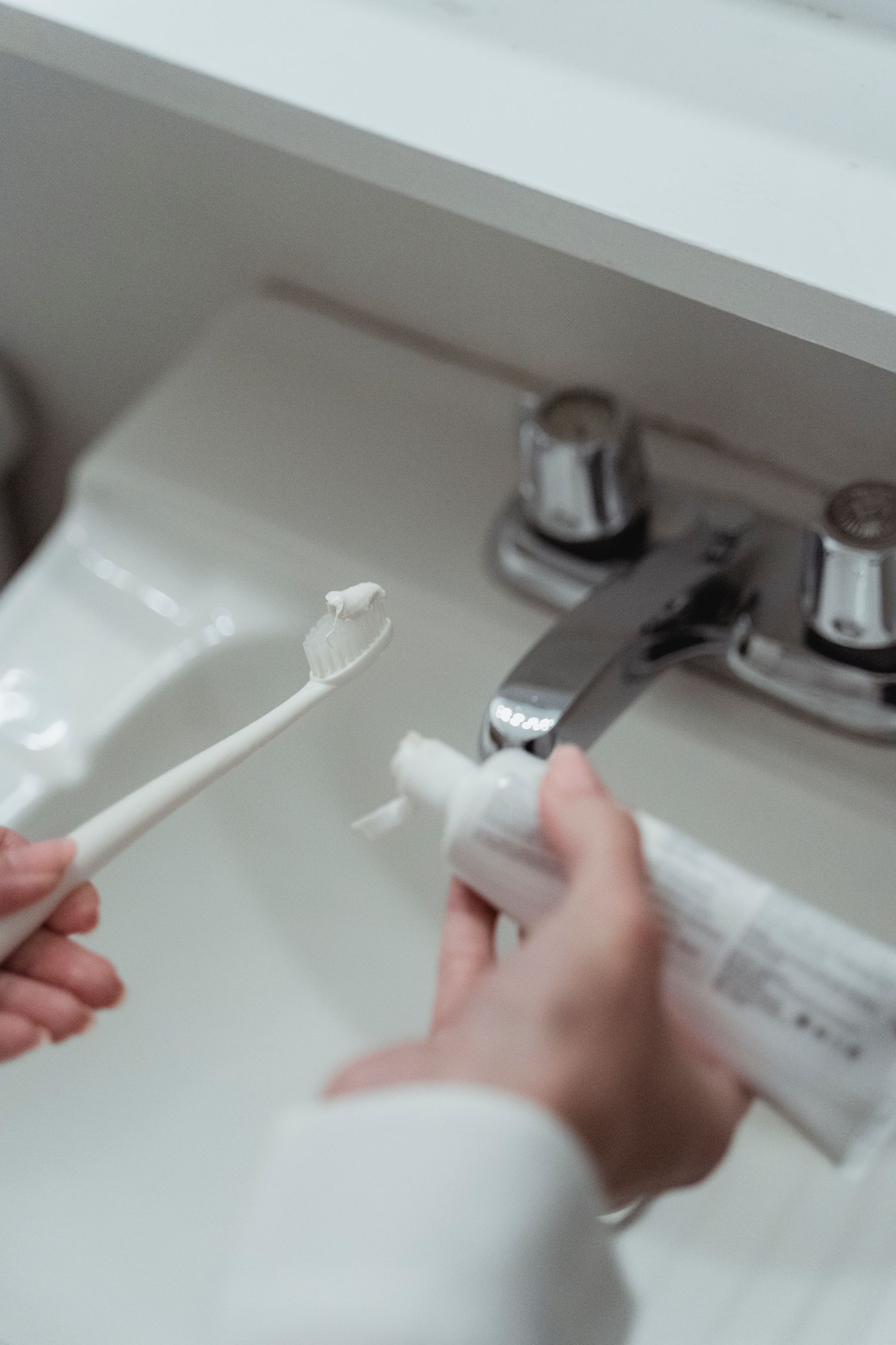 Hands applying toothpaste to a toothbrush above a bright bathroom sink during a morning oral-care routine